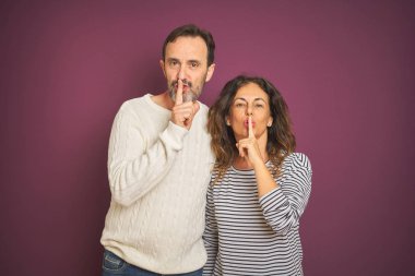 Beautiful middle age couple wearing winter sweater over isolated purple background asking to be quiet with finger on lips. Silence and secret concept. clipart