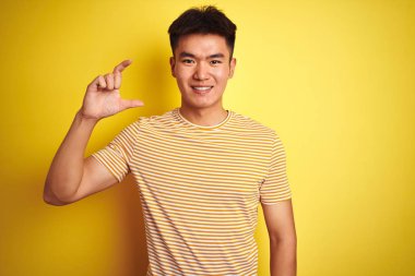 Genç asyalı Çinli adam izole sarı arka plan üzerinde duran t-shirt giyen gülümseyerek ve emin el parmakları görünümlü ve kamera ile küçük boyutlu işareti yapıyor gesturing. Ölçü kavramı.