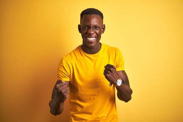 Young african american man wearing casual t-shirt standing over isolated yellow background very happy and excited doing winner gesture with arms raised, smiling and screaming for success. Celebration concept.