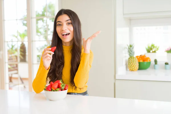 健康的なスナックとして新鮮な赤いイチゴを食べる若い女性は非常に幸せで興奮し 大きな笑顔で叫んで勝利を祝う勝者の表情と手を上げた — ストック写真