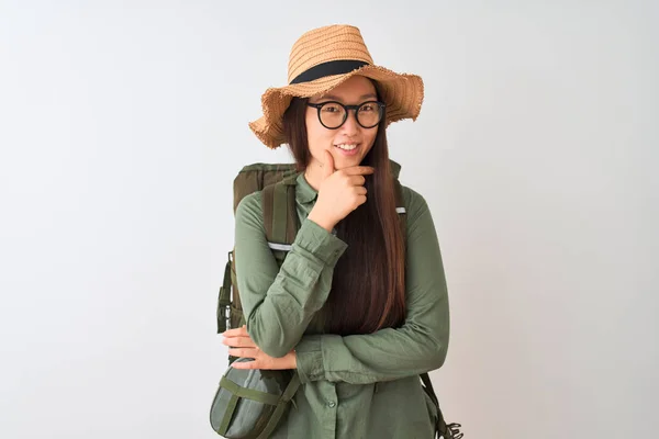 Chinese hiker woman wearing canteen hat glasses backpack over isolated white background looking confident at the camera smiling with crossed arms and hand raised on chin. Thinking positive.