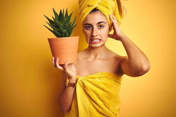 Young beautiful woman wearing a towel holding aloe vera pot over yellow isolated background stressed with hand on head, shocked with shame and surprise face, angry and frustrated. Fear and upset for mistake.
