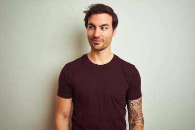 Izole beyaz arka plan üzerinde mor rahat t-shirt giyen dövme ile genç yakışıklı adam yan bakarak ve düşünme uzağa bakıyor.