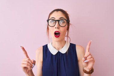 Izole pembe arka plan üzerinde zarif elbise ve gözlük giyen pigtail ile Redhead kadın şaşırttı ve yukarı bakarak ve parmakları ile işaret ve kolları kaldırdı şaşırttı.