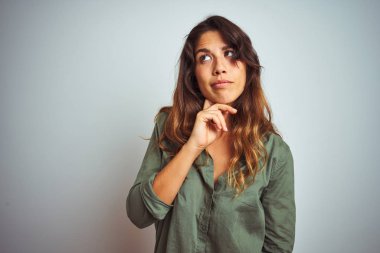 Yeşil gömlek giyen genç güzel kadın, gri izole arka plan üzerinde duran elleri çene üzerinde soru, dalgın ifade düşünme. Düşünceli bir yüzle gülümsüyor. Şüphe kavramı.