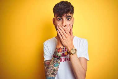 Dövme giyen genç adam izole sarı arka plan üzerinde duran fanny t-shirt giyen hata için elleri ile ağız kapsayan şok. Gizli kavram.