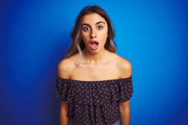 Genç güzel kadın mavi izole arka plan üzerinde çiçek t-shirt giyen korkmuş ve sürpriz ifade, korku ve heyecanlı yüz ile şok.