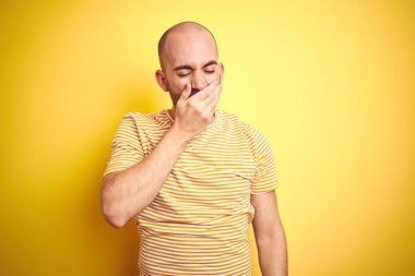 Sakallı genç kel adam sarı izole arka plan üzerinde rahat çizgili t-shirt giyen el ile ağız kapsayan yorgun esneme sıkılmış. Huzursuz ve uykululuk.