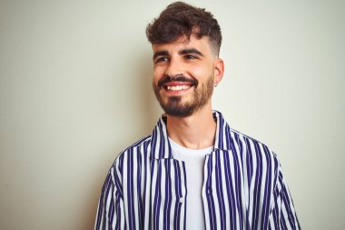Çizgili gömlek giyen dövme giyen genç adam izole beyaz arka plan üzerinde duran yüzünde gülümseme, doğal ifade ile yan bakıyor. Gülmekten emin.