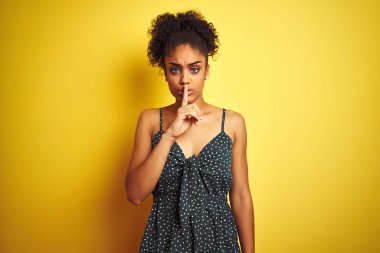 Izole sarı arka plan üzerinde yaz rahat yeşil elbise giyen Afrikalı-Amerikalı kadın dudaklarında parmak ile sessiz olmak isteyen. Sessizlik ve gizli kavram.