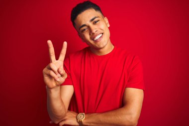 İzole kırmızı arka plan üzerinde duran tişört giyen brezilyalı genç adam mutlu yüzüyle gülümseyerek parmaklarıyla zafer işareti yaparak kameraya göz kırpıyor. İki numara.