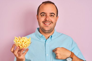 Genç adam kendini işaret parmak işaret sürpriz yüzü ile izole pembe arka plan üzerinde duran ekstrüde mısır ile kase tutan
