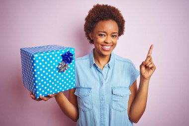 Pembe izole arka plan üzerinde doğum günü hediyesi tutan Afrikalı amerikalı kadın çok mutlu yan el ve parmak ile işaret