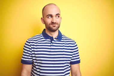 Sakallı genç kel adam sarı izole arka plan üzerinde rahat çizgili mavi t-shirt giyen yan bakarak ve düşünme uzak bakıyor.