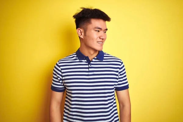 Young asian chinese man wearing striped polo standing over isolated yellow background looking away to side with smile on face, natural expression. Laughing confident.