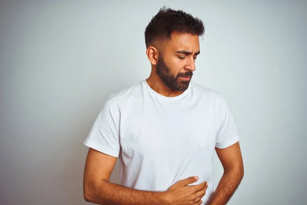 Young indian man wearing t-shirt standing over isolated white background with hand on stomach because indigestion, painful illness feeling unwell. Ache concept.