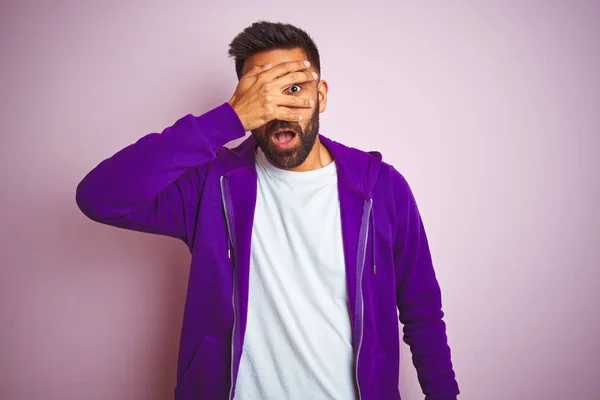 Young indian man wearing purple sweatshirt standing over isolated pink background peeking in shock covering face and eyes with hand, looking through fingers with embarrassed expression.