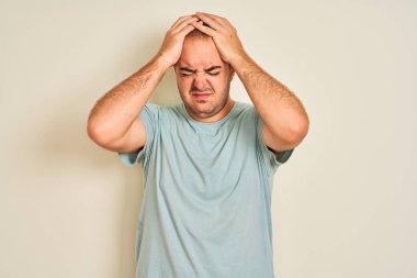 Mavi rahat t-shirt giyen genç adam baş ağrısı umutsuz muzdarip izole beyaz arka plan üzerinde duran ve stresli çünkü ağrı ve migren. Eller baş üstünde.