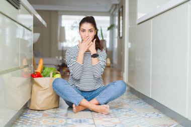 Mutfak zemininde taze bakkaliye dolu bir kese kağıdıyla oturan genç kadın, hata yüzünden ağızlarını elleriyle kapatarak şok emdi. Gizli kavram.