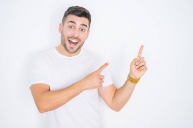 Genç yakışıklı adam beyaz izole arka plan üzerinde rahat beyaz t-shirt giyen gülümseyerek ve yan iki el ve parmakları ile işaret kamera bakarak.