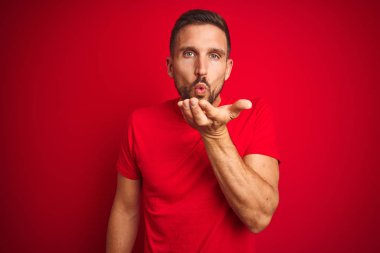 Genç yakışıklı adam kırmızı izole arka plan üzerinde rahat t-shirt giyen kamera hava güzel ve seksi olmak el iyle bir öpücük üfleme bakarak. Aşk ifadesi.