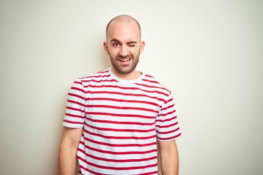 Sakallı genç kel adam beyaz izole arka plan üzerinde rahat çizgili kırmızı t-shirt giyen seksi ifade, neşeli ve mutlu yüz ile kameraya bakarak göz kırpıyor.