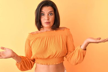 Genç güzel kadın rahat t-shirt izole turuncu arka plan üzerinde duran giyen habersiz ve kolları ve elleri ile karışık ifade kaldırdı. Şüphe kavramı.
