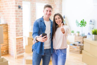 Güzel genç çift ok işareti yapıyor büyük bir gülümseme ile mutlu akıllı telefon kullanarak yeni bir eve hareket, parmakları ile başparmak, mükemmel işareti