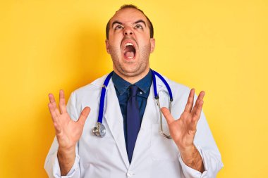 Genç doktor adam ceket ve stetoskop giyen izole sarı arka plan üzerinde duran deli ve deli bağırarak ve agresif ifade ve kollar kaldırdı ile bağırıyor. Hayal kırıklığı kavramı.