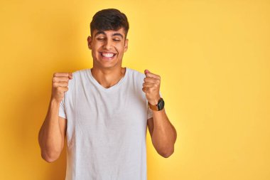 İzole sarı arka plan üzerinde duran beyaz tişört giyen genç hintli adam, kollarını kaldırıp gözleri kapalı zaferi gülümseyerek başarı için heyecanlı. Kazanan kavramı.
