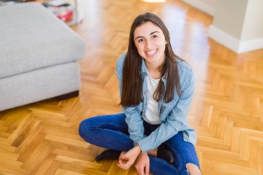 Güzel genç esmer kadın, arka planda güneş ışığı ile evde yerde oturan yüzünde büyük bir gülümseme ile kameraya bakarak neşeli gülümseyen