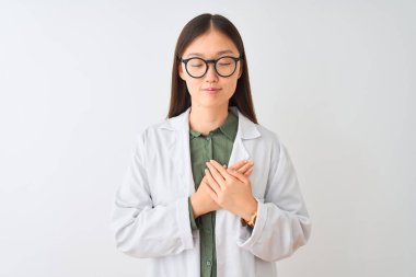 Genç Çinli bilim adamı kadın kapalı gözleri ve yüzünde minnettar jest ile göğüs üzerinde gülümseyerek izole beyaz arka plan üzerinde ceket ve gözlük giyiyor. Sağlık kavramı.