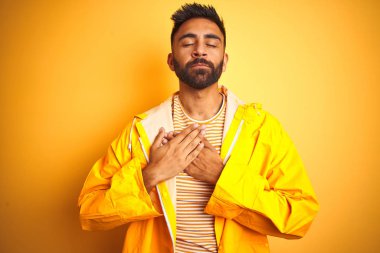 İzole sarı arka plan üzerinde duran yağmurluk giyen genç hintli adam kapalı gözleri ve yüzünde minnettar jest ile göğüs üzerinde gülümseyerek. Sağlık kavramı.