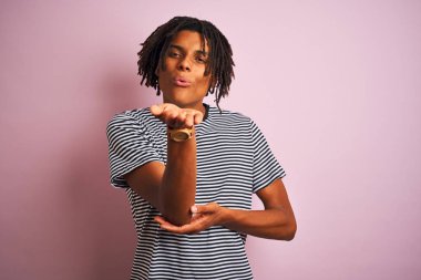 Dreadlocks lacivert çizgili t-shirt giyen afro adam izole pembe arka plan üzerinde duran kamera hava güzel ve seksi olmak el ile bir öpücük üfleme bakarak. Aşk ifadesi.