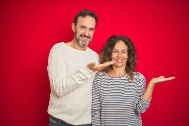 Izole kırmızı arka plan üzerinde kış kazak giyen güzel orta yaş çift neşeli sunan gülümseyerek ve kameraya bakarak avuç içi ile işaret.