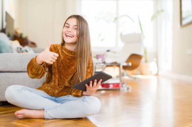 Güzel genç kız çocuk dijital Touchpad tablet kullanarak katta oturan mutlu büyük gülümseme ile ok işareti, başparmak yukarı parmaklar, mükemmel işareti yapıyor