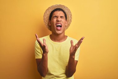 Genç yakışıklı Arap adam izole yelllow arka plan deli ve deli bağırarak ve agresif ifade ve kollar kaldırdı ile bağırıyor üzerinde t-shirt ve yaz şapka giyiyor. Hayal kırıklığı kavramı.