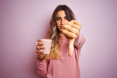 Kızgın yüz ile pembe izole arka plan üzerinde kahve fincan tutan genç güzel kadın, başparmak aşağı, ret kavramı ile sevmediğimi gösteren negatif işareti
