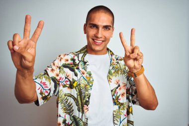 Tatilde beyaz arka plan üzerinde Hawaii gömleği giyen genç yakışıklı adam gülümseyerek kameraya bakarak parmaklarını zafer işareti yapıyor gösteren. İki numara.