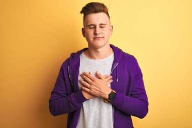 Mor sweatshirt giyen genç yakışıklı adam kapalı gözleri ve yüzünde minnettar jest ile göğüs üzerinde gülümseyerek izole sarı arka plan üzerinde duran. Sağlık kavramı.