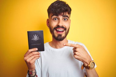Izole sarı arka plan üzerinde Avustralya Avustralya pasaportu giyen dövme ile genç adam çok mutlu el ve parmak ile işaret