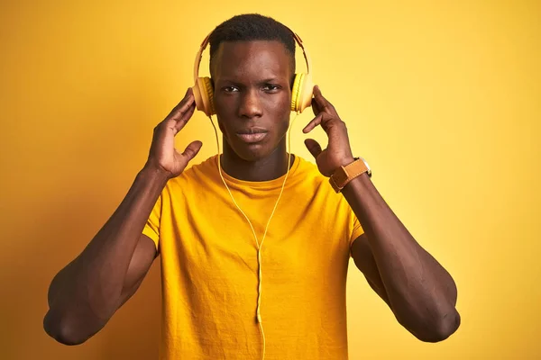 African american man listening to music using headphones over isolated yellow background with a confident expression on smart face thinking serious