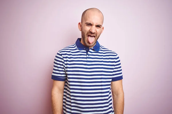 Young bald man with beard wearing casual striped blue t-shirt over pink isolated background sticking tongue out happy with funny expression. Emotion concept.