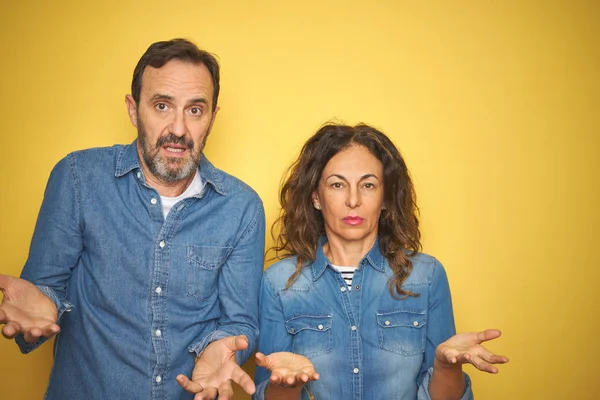 Güzel orta yaş çift birlikte izole sarı arka plan clueless üzerinde duran ve açık kollar ile karışık, hiçbir fikir kavramı.