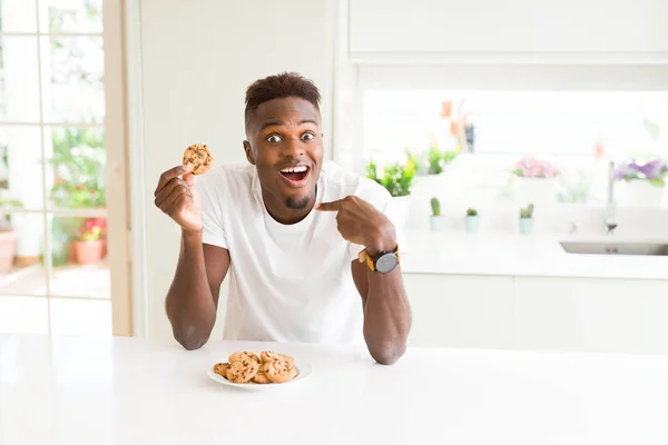 若いアフリカ系アメリカ人男性がチョコレートチップクッキーを食べて驚きの顔で自分自身に指を向ける — ストック写真