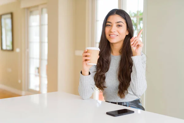 Young woman drinking a cup of coffee at home surprised with an idea or question pointing finger with happy face, number one