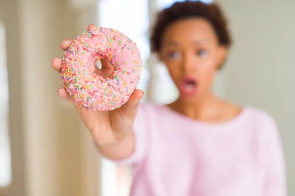 ピンクの砂糖ドーナツを食べる若いアフリカ系アメリカ人女性は 驚きの顔でショックを受け 恐怖表現で恐怖と興奮 — ストック写真