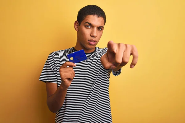 Young handsome arab man holding credit card standing over isolated yellow background pointing with finger to the camera and to you, hand sign, positive and confident gesture from the front