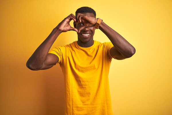 Young african american man wearing casual t-shirt standing over isolated yellow background Doing heart shape with hand and fingers smiling looking through sign