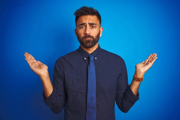 Young indian businessman wearing elegant shirt and tie standing over isolated blue background clueless and confused expression with arms and hands raised. Doubt concept.